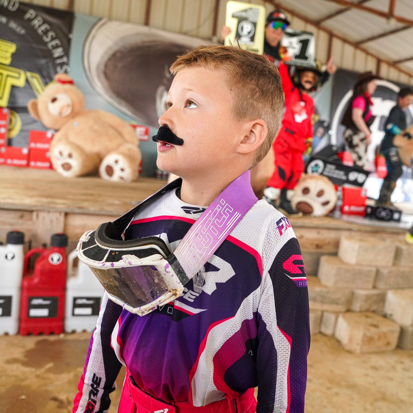 Child in Clutch motocross gear with a mustache and helmet, standing in a indoor setting with various items and people in the background.